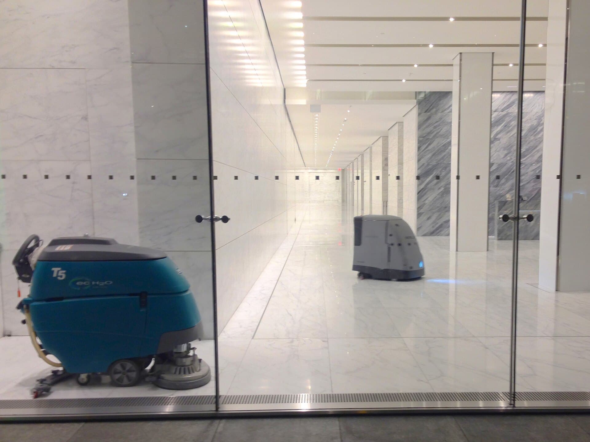 A walk-behind floor scrubber beside an autonomous floor-scrubbing robot