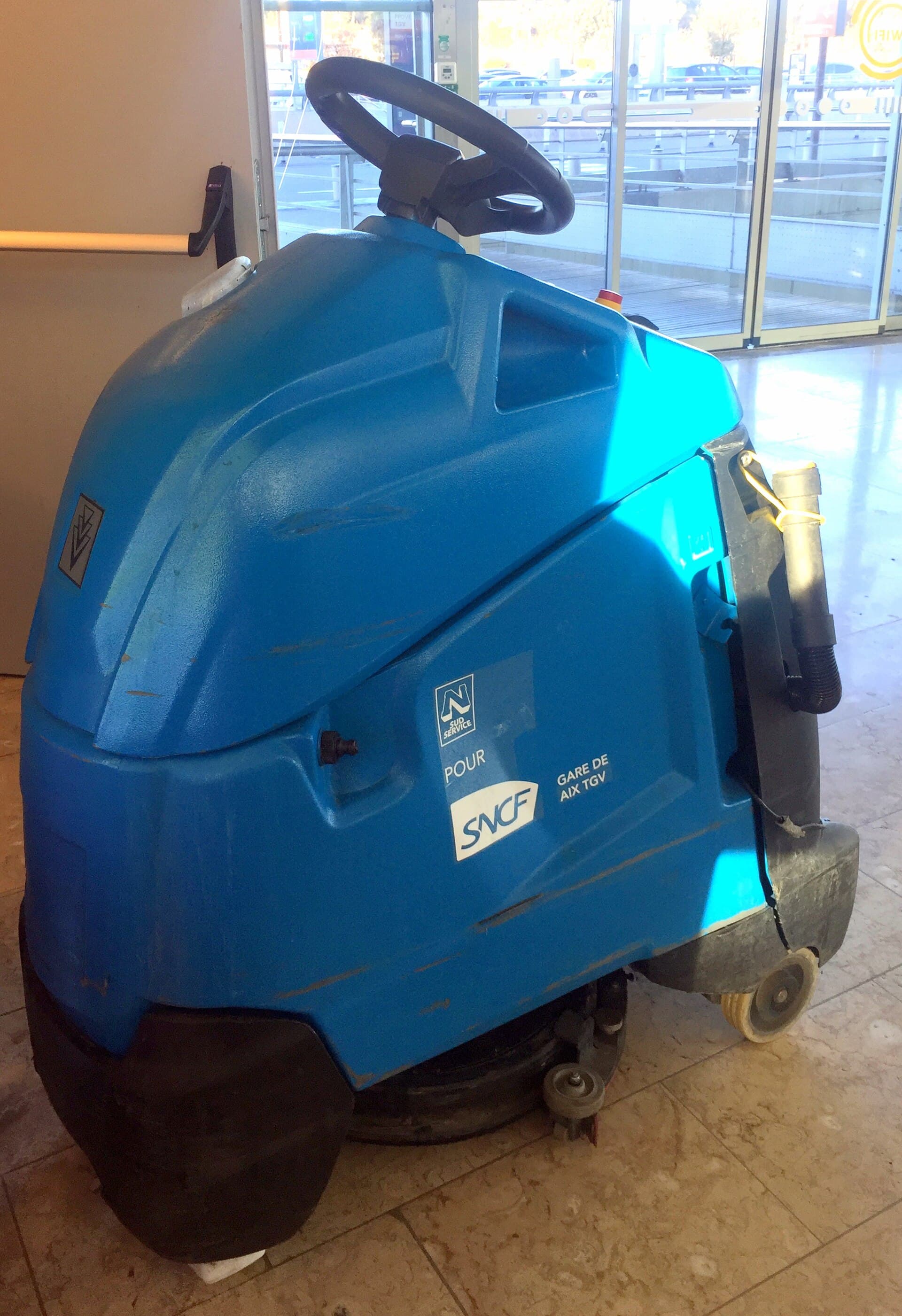 A blue Kärcher walk-behind scrubber-dryer parked on a tiled concourse